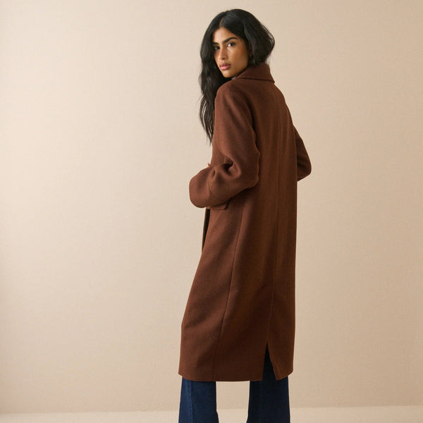 A woman modeling a long brown wool-blend coat, shown from a three-quarters back view. She is wearing blue wide-leg jeans and dark brown heeled boots against a solid beige studio background.