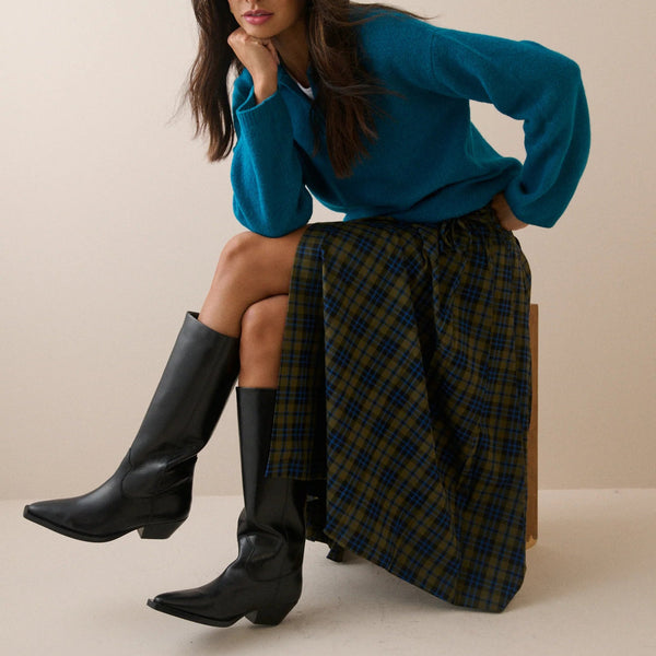 A woman models a teal knit sweater and a green and navy plaid skirt while sitting on a wooden stool. She is wearing black knee-high leather boots against a solid beige studio background.