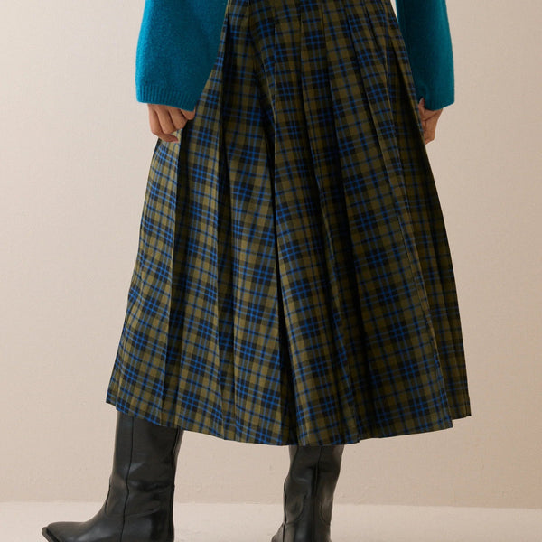 A pleated A-line skirt in a blue and olive green tartan plaid pattern, styled with a teal sweater and black leather ankle boots, shown from the back against a neutral beige background.