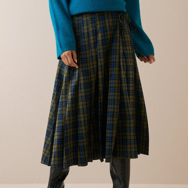 A person wearing a pleated A-line skirt in a green and blue tartan plaid pattern, styled with a teal sweater and black leather boots against a neutral beige background.