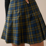 A close-up of a pleated plaid skirt in olive green, blue, and black, worn by a model with a black top visible at the waist.