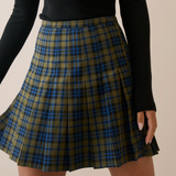 A pleated plaid skirt in olive green, black, and blue, worn by a model in a black long-sleeve top against a plain beige background.