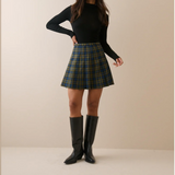 A woman stands against a plain beige studio background wearing a fitted black long-sleeve top, a green and navy plaid mini skirt, and black knee-high boots.