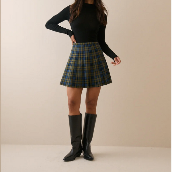 A woman stands against a plain beige studio background wearing a fitted black long-sleeve top, a green and navy plaid mini skirt, and black knee-high boots.