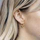 A close-up of a person's ear wearing a small textured gold hoop earring, with blonde hair framing the side of the face.