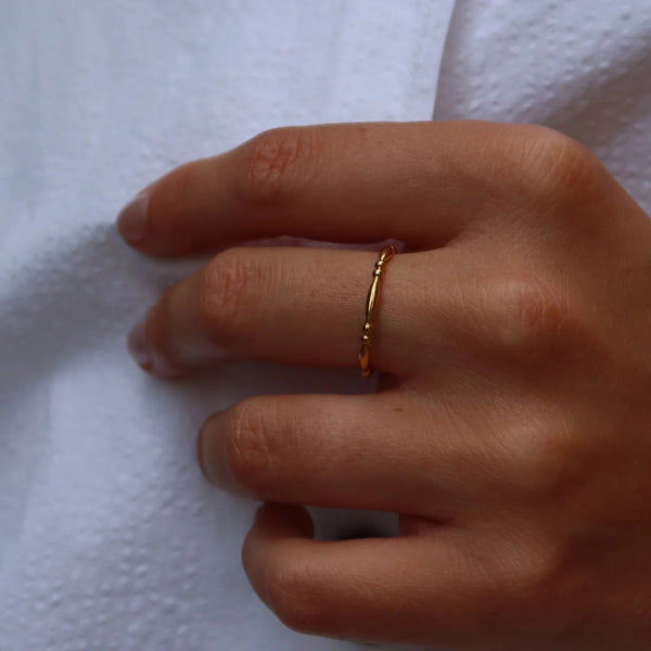 A close-up of a person's hand wearing a thin gold ring on one of the middle fingers, set against a white textured fabric background.