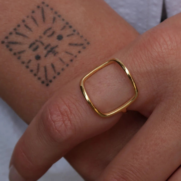 A close-up of a hand wearing a thin gold ring with a square openwork design, with a black geometric tattoo visible on the adjacent wrist.