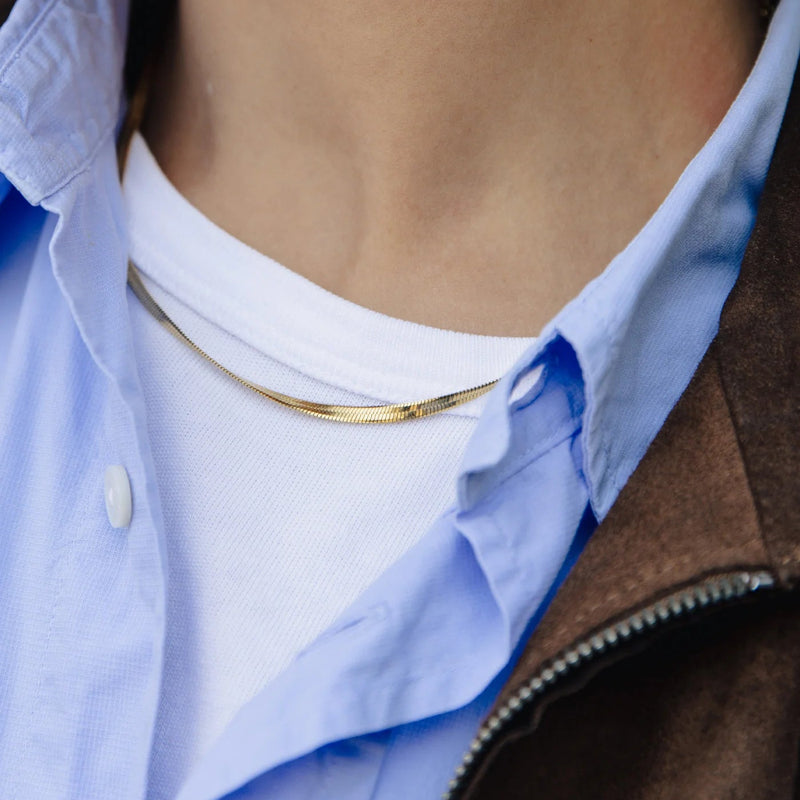 Close-up of a person wearing a gold necklace with a blue shirt and brown jacket.