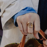 Hand wearing a gold ring holding a brown leather strap, with a blurred background.