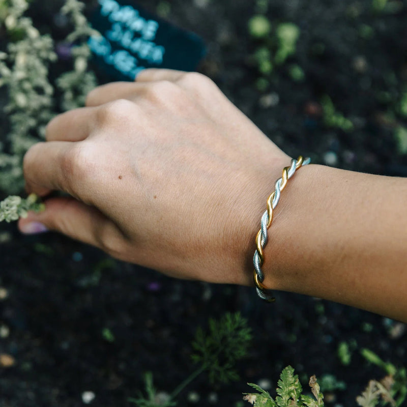 A close-up of a person's hand and wrist wearing a twisted metal bracelet outdoors, with dark soil and small green plants in the background.