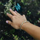 A close-up of a person's hand wearing a twisted gold and silver bangle bracelet, resting near small green plants in dark soil.