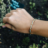 A close-up of a person's hand and wrist wearing a twisted metal bracelet outdoors, with dark soil and small green plants in the background.