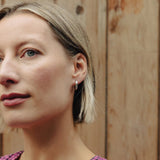 A close-up portrait of a woman with short blonde hair wearing a pink and black gingham patterned top, set against a rustic wooden plank background.