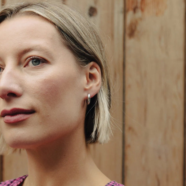 A close-up portrait of a woman with short blonde hair wearing a pink and black gingham patterned top, set against a rustic wooden plank background.