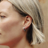 A woman in profile wearing a small silver huggie hoop earring, with blonde hair and a pink patterned top against a wooden background.