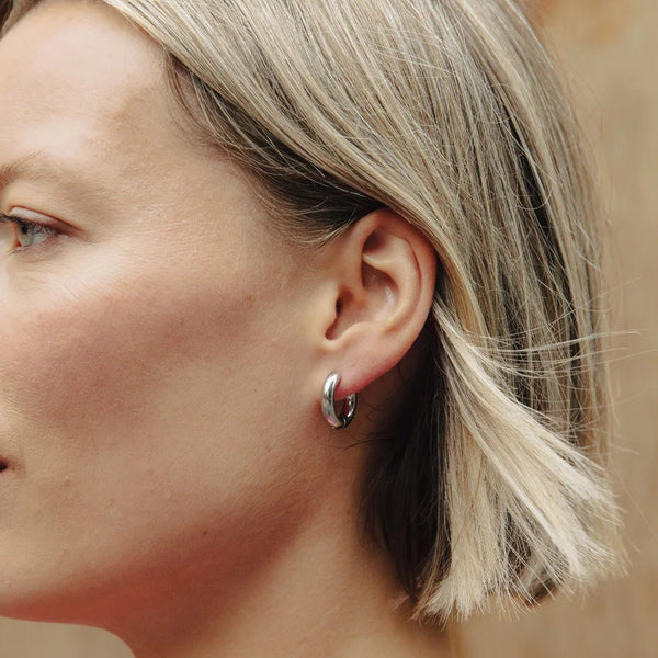 A woman in profile wearing a small silver huggie hoop earring, with blonde hair and a pink patterned top against a wooden background.