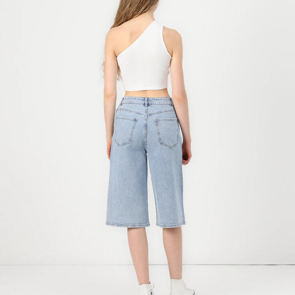 A model shown from the back wearing light-wash blue cropped denim shorts, a white one-shoulder crop top, and white sneakers against a plain white studio background.