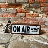 A rectangular vintage-style 'ON AIR' sign with a white face, black lettering, and a brown wooden frame, placed on a stone surface against a red brick wall. The sign reads 'ON AIR' with 'BBC STUDIO' printed on it, and a black power cord is visible on the left.