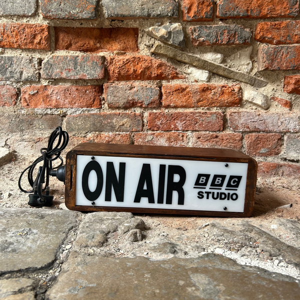 A rectangular vintage-style 'ON AIR' sign with a white face, black lettering, and a brown wooden frame, placed on a stone surface against a red brick wall. The sign reads 'ON AIR' with 'BBC STUDIO' printed on it, and a black power cord is visible on the left.