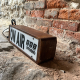 A rectangular wooden sign with a white face reading 'ON AIR BBC STUDIO' in black text, photographed resting on a dirt and stone surface against a red brick wall.