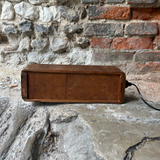 A vintage rectangular wooden box with a power cord plugged in, resting on a stone surface against a rustic brick and stone wall background.
