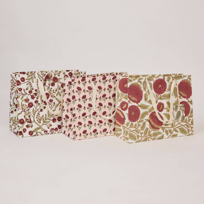 Three fabric gift bags with botanical patterns in red, green, and beige, arranged in a row against a plain light gray background.