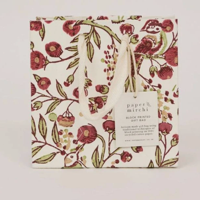 A Paper Mirchi block-printed gift bag featuring a botanical pattern of red flowers, green leaves, and small birds on a cream background, with white rope handles and a white brand tag.