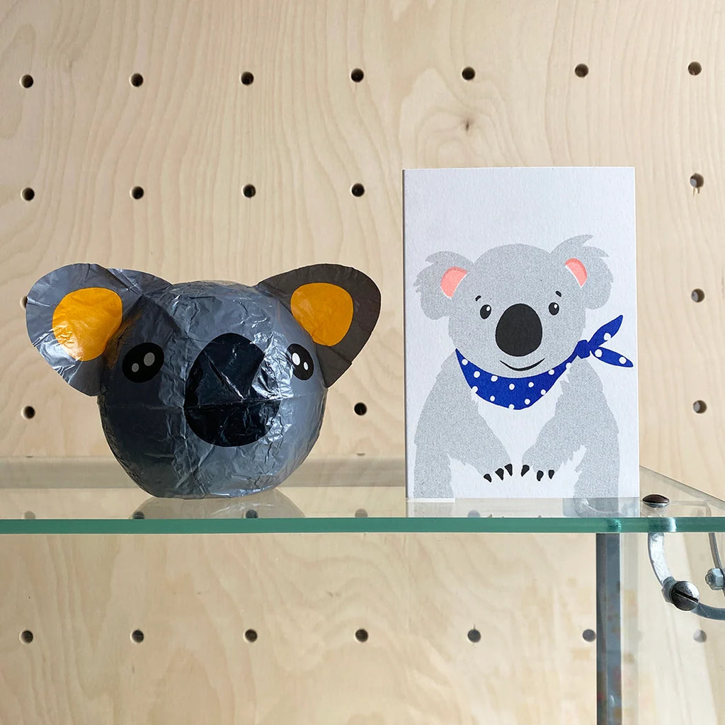 Model of a koala head and a card with a koala design on a glass shelf against a wooden background.