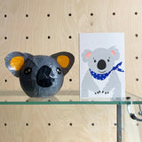 Model of a koala head and a card with a koala design on a glass shelf against a wooden background.