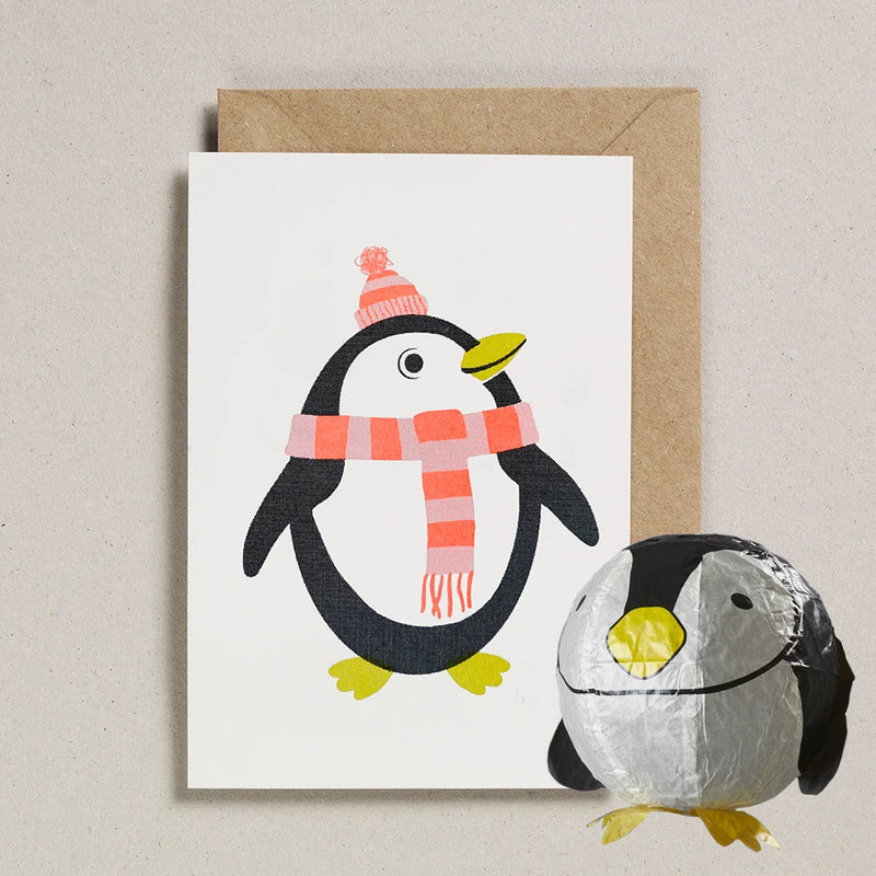 A white greeting card featuring a cartoon penguin wearing a pink and orange striped hat and scarf, shown with a brown kraft envelope behind it and a small foil-wrapped puffin toy in front on a light beige textured background.