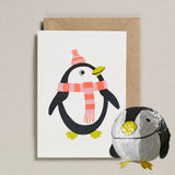 A white greeting card featuring a cartoon penguin wearing a pink and orange striped hat and scarf, shown with a brown kraft envelope behind it and a small foil-wrapped puffin toy in front on a light beige textured background.