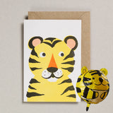 A children's greeting card featuring a yellow tiger illustration with black stripes, shown with a brown kraft envelope and a small matching yellow tiger balloon on a light gray background.