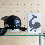 Black paper mache orca whale sculpture next to a card with a whale design on a wooden surface.