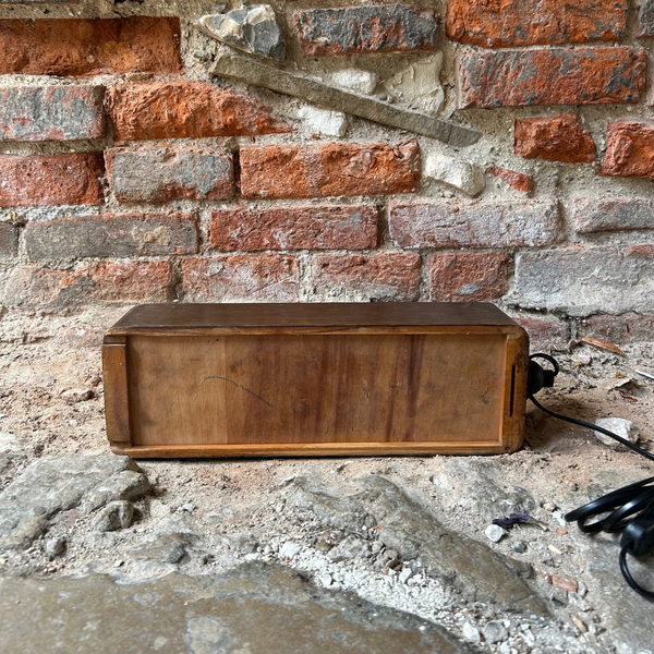 A rectangular wooden chest or speaker cabinet with a dark brown wood finish sits on a rough concrete floor against a red and gray brick wall. A black power cord is visible on the right side.