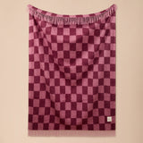 A pink and purple checkerboard patterned throw blanket with fringe on the top and bottom, hanging against a solid beige wall.