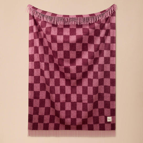 A pink and purple checkerboard patterned throw blanket with fringe on the top and bottom, hanging against a solid beige wall.