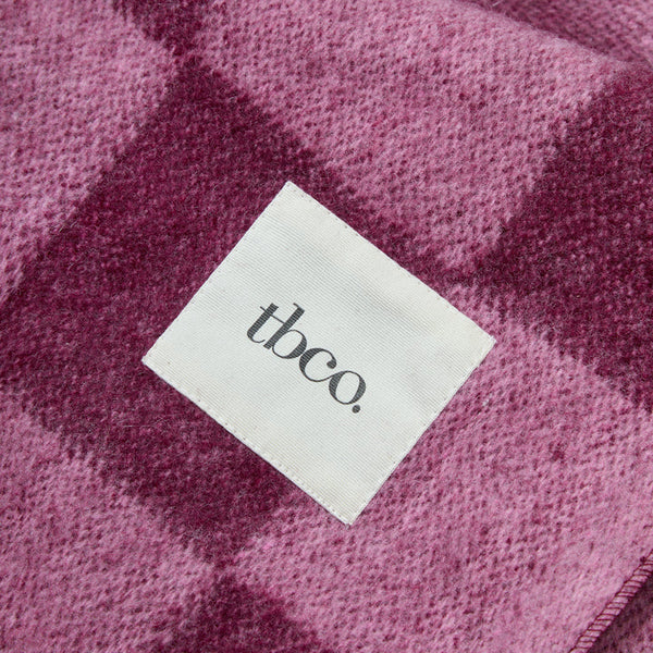A close-up of a textured woven blanket in a dusty mauve and dark plum checkered pattern, featuring a white square label with the brand name 'tbco.' printed in black.