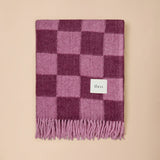 A folded checkered scarf in shades of pink and purple with a small white brand label reading 'the co.' and fringed edges, displayed against a solid beige background.