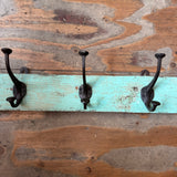 Three black metal coat hooks mounted on a distressed turquoise painted wooden board, set against a rustic brown wood plank background.