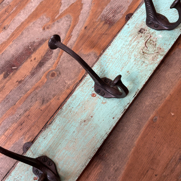 A rustic three-hook coat rack made from a distressed light blue painted wooden plank with dark metal hooks, shown on a warm brown wood floor.