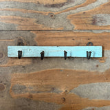 A rustic light blue wooden wall hook rack with four black metal hooks, mounted on a weathered wood plank wall.
