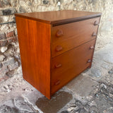 Stag mid-century modern three-drawer wooden chest of drawers with a warm reddish-brown finish and dark round knobs, set against a rustic stone wall and stone floor.