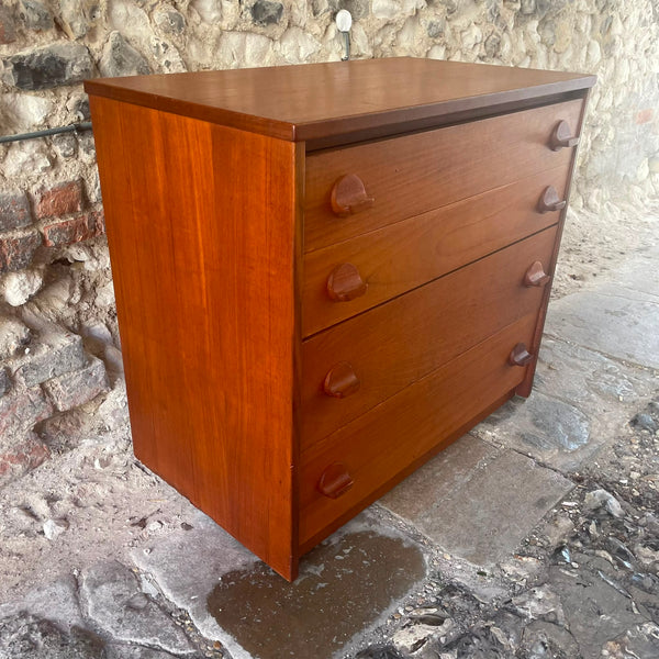 Stag mid-century modern three-drawer wooden chest of drawers with a warm reddish-brown finish and dark round knobs, set against a rustic stone wall and stone floor.