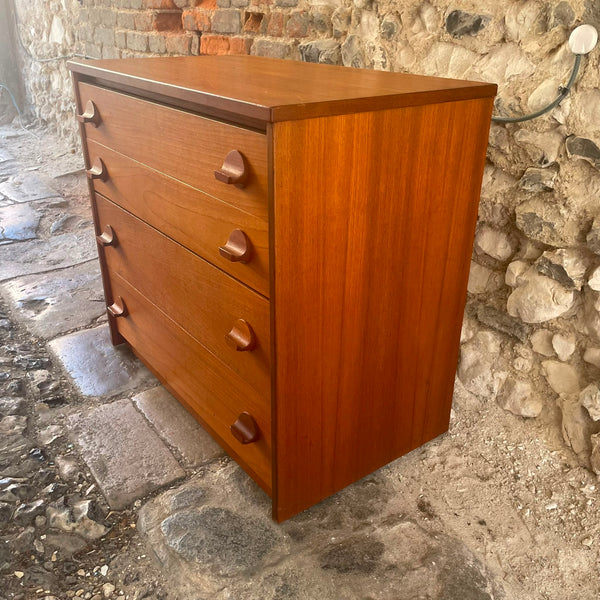 Stag mid-century modern wooden chest of drawers with four drawers and round knobs, made of warm reddish-brown wood, photographed outdoors against a stone wall.