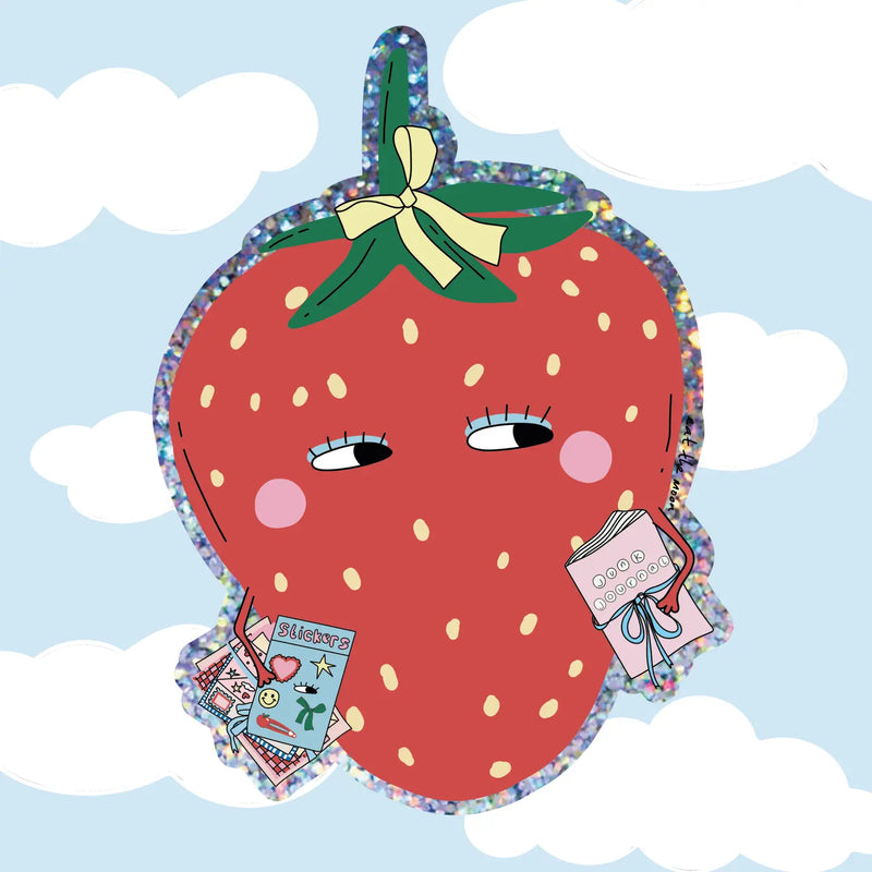 A strawberry-shaped plush or decorative item with a glittery blue border, cartoon eyes, rosy cheeks, a green leafy top with a yellow bow, and small attached tags, shown against a light blue sky with white clouds.