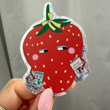 A hand with pink manicured nails holds a die-cut sticker shaped like a red strawberry. The sticker features a glossy red body with yellow seeds, a green leafy top with a yellow bow, and a holographic silver border.