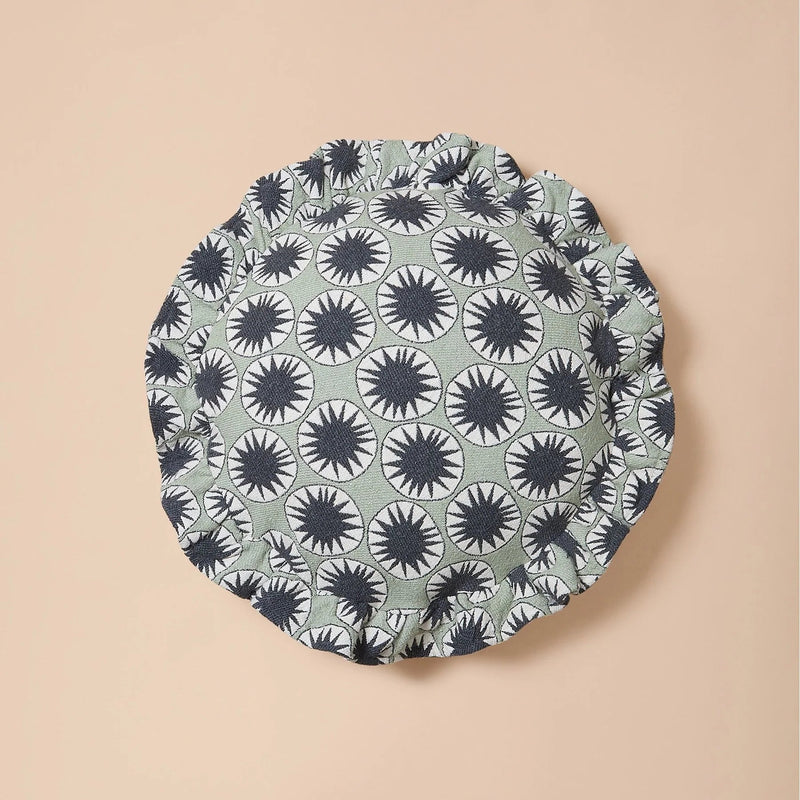 A round fabric headband or bonnet with a repeating black and white sunburst pattern on a muted sage green background, displayed flat against a solid beige backdrop.