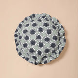 A round fabric headband or bonnet with a repeating black and white sunburst pattern on a muted sage green background, displayed flat against a solid beige backdrop.