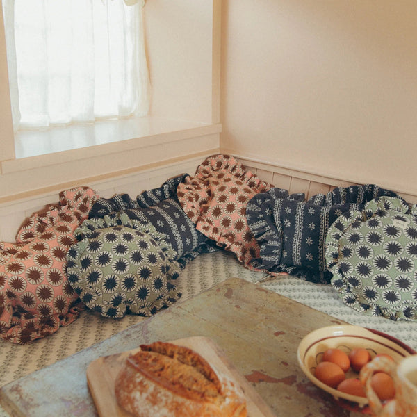 A cozy corner of a room with a built-in bench cushioned in patterned pillows, with a rustic table in the foreground holding a loaf of bread and a bowl of eggs.