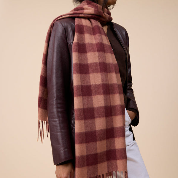 A woman wearing a large brown and burgundy plaid scarf with fringe, styled over a dark brown leather jacket and white pants, set against a solid beige studio background.